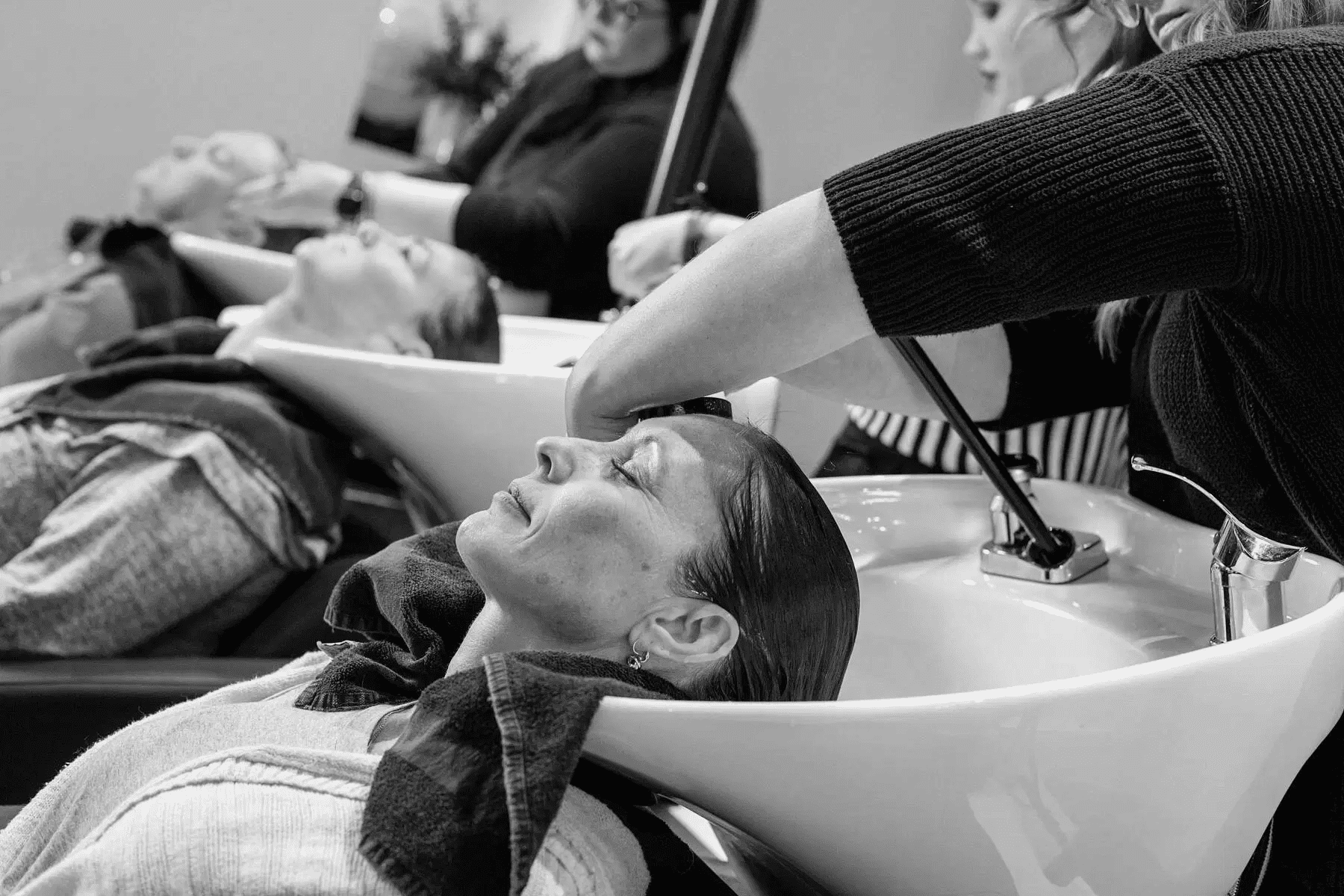 People getting their hair washed at a salon sink, attended by stylists.